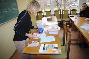 Zdjęcie na sali szkoleniowej - jedna z koordynatorek przedsięwzięcia przygotowuje materiały dla uczestników a w tle widać jednego ze słuchaczy siedzących za stołem