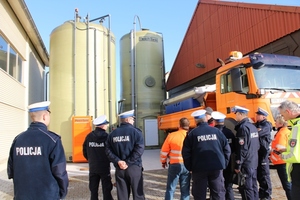 Zdjęcie grupy uczestników szkolenia (ubrani w granatowe mundury - dwie osoby mają na sobie kamizelki pomarańczowe i jedna osoba - żółtą) - stojący w plenerze (ujęcie od tyłu - widoczne budynki oraz dwa pionowe zbiorniki i pomarańczowy samochód ciężarowy)