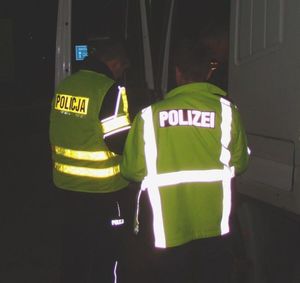 Zdjęcie dwóch funkcjonariuszy - ujęcie w nocy od tyłu - jeden z policjantów ma na plecach kamizelki odblaskowej napis "Policja" a drugi - "Polizei"