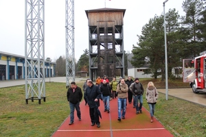 Zdjęcie grupowe uczestników na tle koszar strażackich - ujęcie od frontu - po lewej stronie widoczny hangar - w środku widoczna drewniana wieża obserwacyjna - po prawej stronie widoczny wóz bojowy
