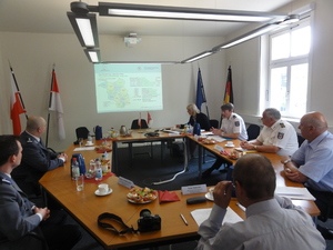 Zdjęcie uczestników spotkania uzgodnieniowego w trakcie obrad - wszyscy siedzą za okrągłym stołem, z przodu sali widoczny jest ekran z wyświetloną prezentacją, a po obu jego bokach flagi (polskie - po lewej, unijna i niemiecka - po prawej)