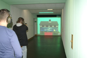 Zdjęcie dwóch uczestników ćwiczeń strzleckich w ochraniaczach - jeden celuje do wizerunku domu wyświetlonego na ekranie