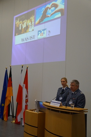 Zdjęcie Insp. Dariusza Banachowicza przy mównicy - obok tłumaczka (obie osoby w mundurach) w tle cztery flagi i ekran z wyświetloną prezentacją