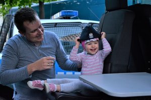 Wnętrze radiowozu policyjnego. Trzyletnia dziewczynka przymierza policyjną furażerkę. Pomaga jej uśmiechnięty ojciec.