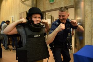 Aula komendy. Wystawa sprzętu policyjnego. Chłopiec ubrany w czarną policyjną kamizelkę i kask pozuje do zdjęcia wraz z policjantem Wydziału Konwojowego.