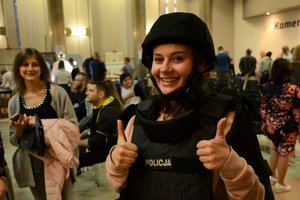 Aula komendy. Wystawa sprzętu policyjnego. Młoda kobieta ubrana w policyjną kamizelkę i kask uśmiecha się i pokazuje kciuki wzniesione do góry.