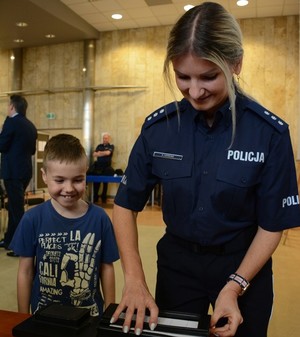 Aula komendy. Wystawa sprzętu policyjnego. Policjantka przygotowuje kartę daktyloskopijną. Obok stoi kilkuletni uśmiechnięty chłopiec.