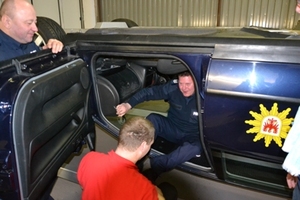 Zdjęcie przedstawiające symulator samochodowy (odwrócony pojazd) - dwóch mężczyzn stojących obok pojazdu i jeden siedzący w kabinie kierowcy