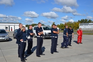 Zdjęcie spośród części uczestników szkolenia stojących w trakcie zajęć na placu manewrowym - jedna osoba w uniformie czerwonym i osiem osób w mundurach granatowych