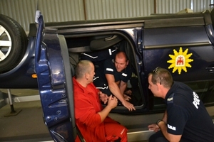 Zdjęcie przedstawiające symulator samochodowy (odwrócony pojazd) - dwóch mężczyzn przykucnięci obok pojazdu i jeden siedzący w kabinie kierowcy