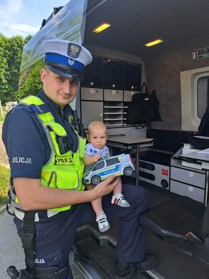 Policjant Ruchu Drogowego trzyma na ręku dziecko. W tle radiowóz policyjny.
