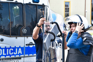Policjant oddziału prewencji policji pomagający przymierzyć kobiecie kamizelkę przeciwuderzeniową, tarczę i pałkę policyjną. W tle oznakowany radiowóz