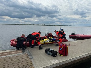 Na zdjęciu widać łódź straży pożarnej oraz wodnego ochotniczego pogotowia ratunkowego. Przeprowadzana jest akcja ratunkowa przez dwóch strażaków oraz trzech ratowników medycznych, wobec osoby poszkodowanej, która znajduje się w łodzi.