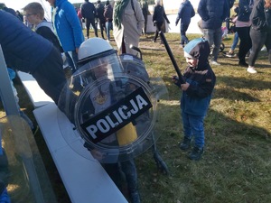 Dzieci przymierzają wyposażenie policjanta prewencji, biorącego udział w zabezpieczeniach meczów piłkarskich i zgromadzeń. W tle tłum ludzi, biorących udział w wydarzeniu.