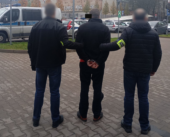 Policjanci trzymają mężczyznę, który ma kajdanki założone na ręce z tyłu.