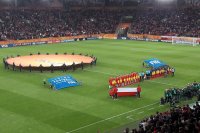 Otwarcie Mistrzostw Świata U-20. Stadion Miejski w Łodzi. Na murawie stoją drużyny piłkarskie Polski i Kolumbii. Za nimi grupa kilkudziesięciu osób trzyma okrągły baner z logo mistrzostw.