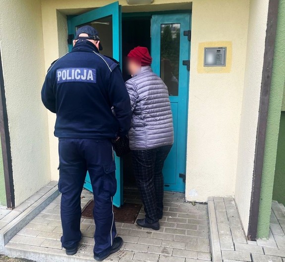 Policjant stoi przed klatką wejściową do budynku wielorodzinnego z zagubioną seniorką.