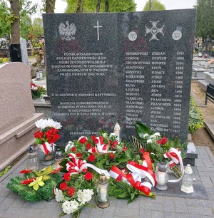 Tablica pamiątkowa z wypisanymi nazwiskami pomordowanych policjantów. Pod płyta leżą kwiaty i pala się znicze.