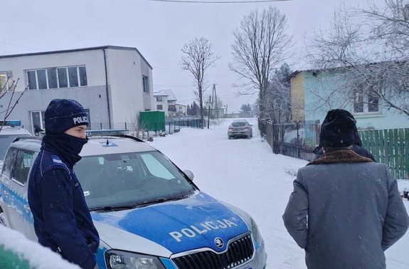 radiowóz, policjant, osoba w kurtce i czpace, budynki.