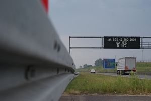 Zdjęcie pobocza autostrady w zbliżeniu - widoczna barierka ochronna oraz trawnik a także umieszczona nad autostradą tablica z wyświetlanymi dla kierowców informacjami