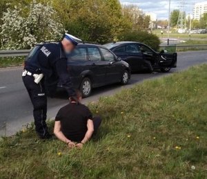 Policjant stoi przy siedzącym na trawie zatrzymanym.