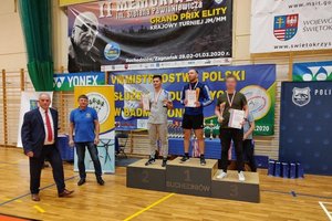 Wnętrze sali gimnastycznej, na podium stoi trzech zawodników, w rękach trzymają medale i dyplomy, po lewej stronie stoi dwóch przedstawicieli komisji zawodów, w tle plakaty i banery.