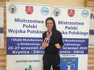 Zawodniczka (brunetka w kitce, ubrana na czarno) z medalem na tle banerów o mistrzostwach.