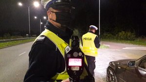 Umundurowany policjant w białej czapce i w maseczce stoi w nocy na drodze i pokazuje wyświetlacz radaru, który pokazuje 142 km, w tle inny policjant przy aucie.