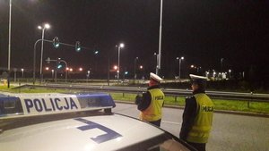 dwaj umundurowani policjanci w białych czapkach i kamizelkach odblaskowych stoja przy drodze, widac dach radiowozu, jest ciemno
