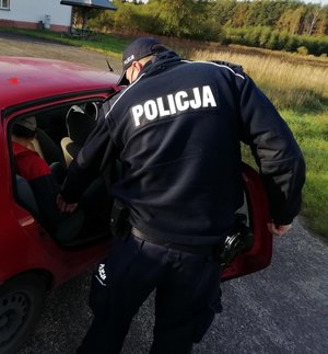 Policjant w granatowym mundurze z białym napisem policja na plecach nachyla się do siedzącego w aucie pasażera.