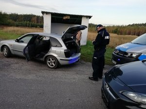 Murowany przystanek autobusowy przed którym stoi otwarte auto a przy nim umundurowany policjant.