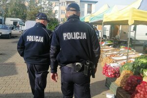 Targowisko miejskie, umundurowani policjanci patrolują teren, w tle stragany handlowe.
