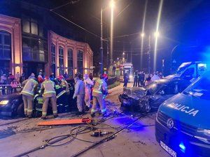 Strażacy i medycy stoją przy rozbitych samochodach i udzielają pomocy osobie zakleszczonej w czarnym samochodzie. Wokół radiowozy policyjne, tramwaj i okoliczni mieszkańcy.