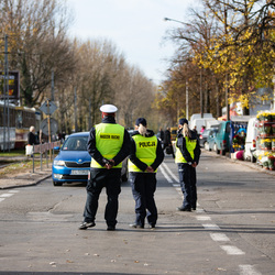 Troje umundurowanych policjantów kieruje ruchem  na ulicy przed cmentarzem.