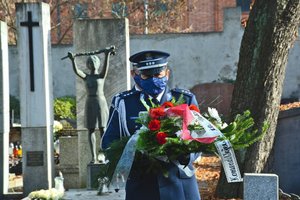 Policjant w mundurze galowym niesie symboliczny wieniec pomiędzy nagrobkami starego cmentarza.