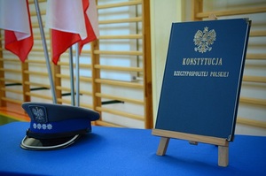 Stół pokryty niebieskim suknem, po lewej stronie oficerska czapka policyjna, po prawej stronie Konstytucja Rzeczypospolitej Polskiej na drewnianym stojaku.