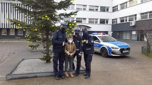 Dwie policjantki i policjant oraz chłopiec stoją przed choinka, która ma odblaskowe elementy, w tle radiowóz.