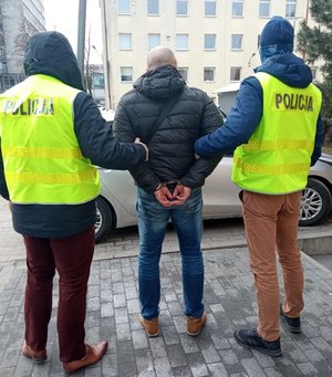 Dwaj policjanci w kamizelkach odblaskowych  trzymają po środku zatrzymanego w czarnej kurtce sportowej z kapturem i jeansach. w tle budynek komendy i nieozakowany radiowóz.