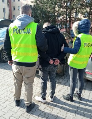 Dwoje policjantów w kamizelkach odblaskowych z napisem policja prowadzą zatrzymanego.