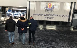 Nieumundurowani policjanci konwojują podejrzanego o przestępstwo korupcyjne. Mężczyzna ma założone kajdanki, w tle znajduję się napis Komenda Wojewódzka Policji w Łodzi.