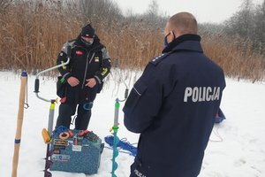 policjant z wędkarzem stoją na oblodzonym zbiorniku wodnym. w tle sprzęt poprawiający bezpieczeństwo na lodzie,
