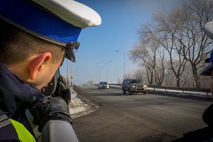 Policjant w białej czapce stoi przy drodze i radarem mierzy prędkość pojazdów.
