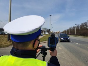 Policjant w białej czapce stoi przy drodze z radarem i mierzy prędkość nadjeżdżającego auta.