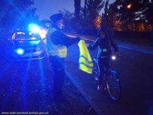 w nocy policyjny radiowóz przy drodze i policjant, który daje rowerzystce kamizelkę odblaskową