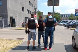 Na dziedzińcu Komendy Miejskiej Policji w Łodzi dwóch policjantów prowadzi zatrzymanego mężczyznę.