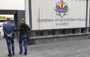 Dziedziniec komendy z tablica  z napisem Komenda Wojewódzka Policji w Łodzi, policjant po cywilnemu prowadzi zatrzymanego.