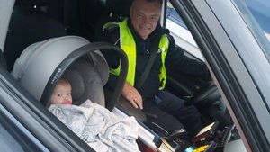 Na zdjęciu widać wnętrze nieoznakowanego radiowozu wyposażonego w videorejstrator.
Za kierownicą siedzi umundurowany policjant ruchu drogowego, na przednim fotelu pasażera w nosidełku uśmiechnięty niemowlak.