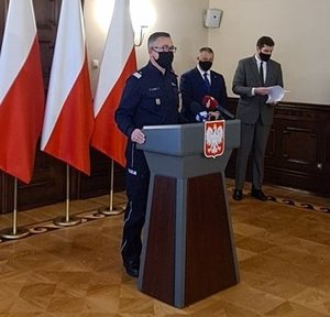 Generał policji stoi za mównicą i zabiera głos, w tle dwóch mężczyzn tj. wojewoda i kurator.obok trzy flagi biało-czerwone.