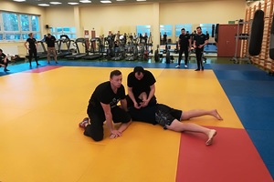 Sala gimnastyczna, zajęcia na macie, policjanci ćwiczą taktyki i techniki interwencji pod okiem instruktorów.