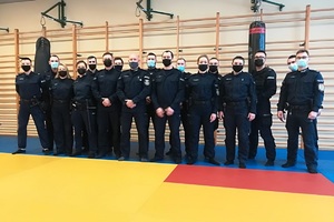 Sala gimnastyczna, zajęcia na macie, policjanci ćwiczą taktyki i techniki interwencji pod okiem instruktorów.
